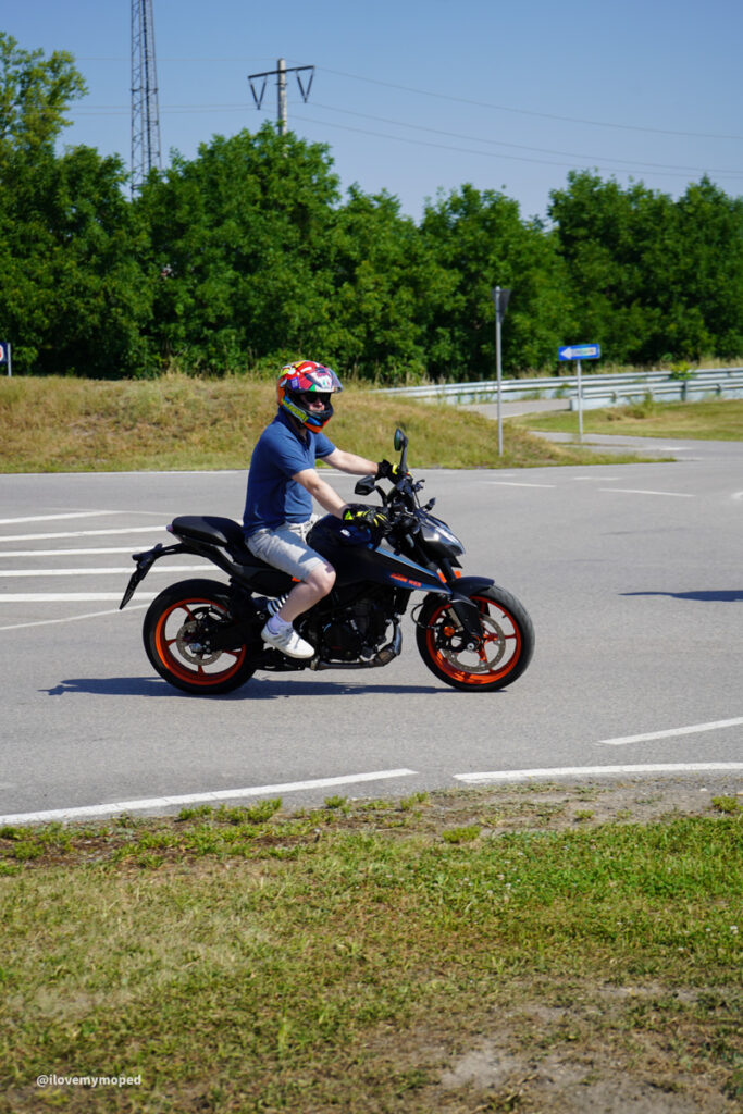 arge2rad-125ertesttag-arböfahrsicherheitszentrumwien-020 - I love my moped Motorcyclist wearing a helmet and blue T‑shirt rides a black sport bike with orange wheels through a roundabout area. Note: concise informative description for a riding scene with proper context and function obvious to screen readers.