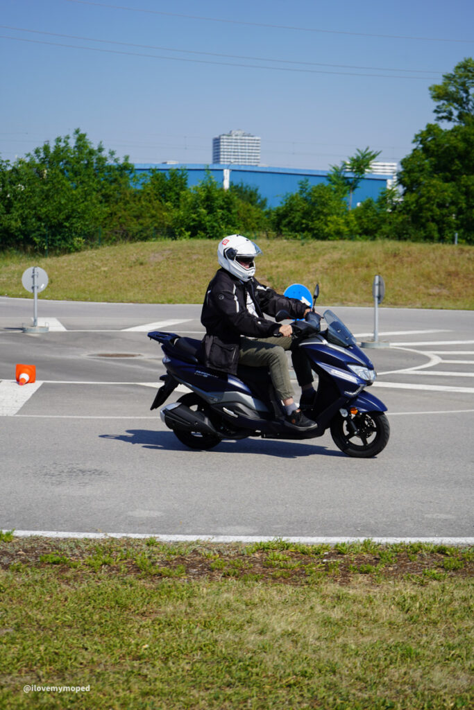 arge2rad-125ertesttag-arböfahrsicherheitszentrumwien-021 - I love my moped A scooter rider wearing a white helmet and black jacket rides a blue scooter on a paved road near a roundabout, with greenery and buildings in the background.