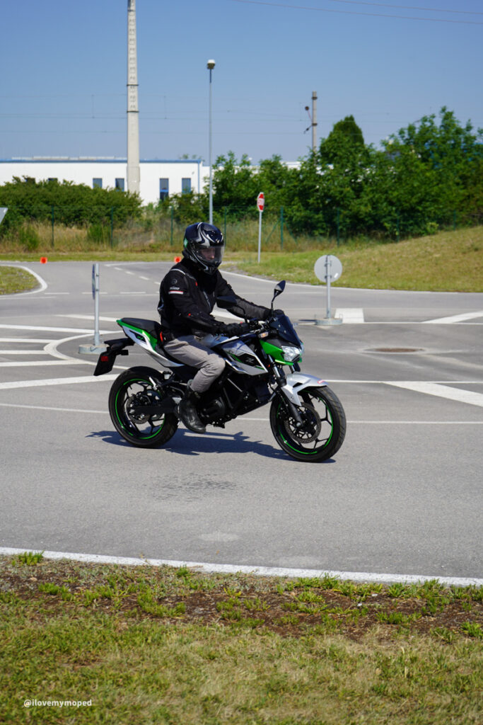 arge2rad-125ertesttag-arböfahrsicherheitszentrumwien-022 - I love my moped Motorcyclist wearing a black helmet and jacket riding a green-and-black sport bike on a paved roundabout under a clear sky.