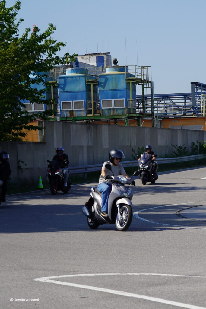 arge2rad-125ertesttag-arböfahrsicherheitszentrumwien-023 - I love my moped Riders on scooters navigate a curved road on a sunny day, with an industrial complex and large blue tanks in the background.