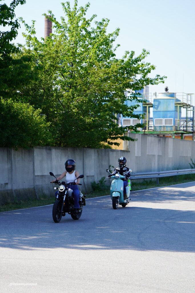 arge2rad-125ertesttag-arböfahrsicherheitszentrumwien-024 - I love my moped Two riders on a black motorcycle and a turquoise scooter ride along a sunlit road beside a concrete barrier, with trees and industrial buildings in the background.