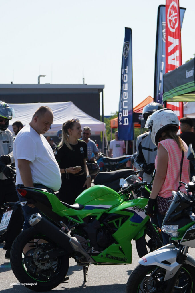arge2rad-125ertesttag-arböfahrsicherheitszentrumwien-025 - I love my moped Green Kawasaki Ninja motorcycle on display at an outdoor event, surrounded by people and tall sponsor banners; a man in a white shirt and others examine the bikes.