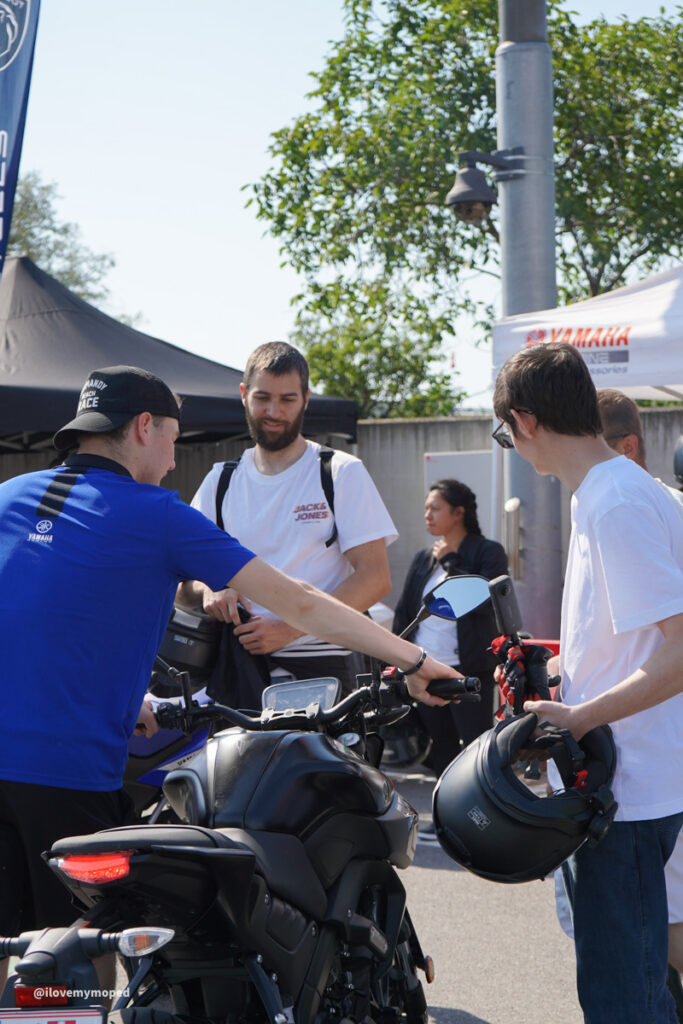 arge2rad-125ertesttag-arböfahrsicherheitszentrumwien-028 - I love my moped Group of people at an outdoor motorcycle event around a black Yamaha bike, one man points at the handlebars.