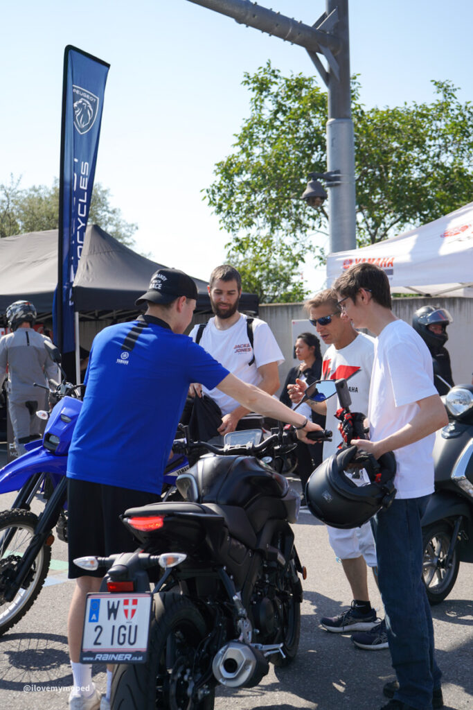 arge2rad-125ertesttag-arböfahrsicherheitszentrumwien-029 - I love my moped Group of men at a motorcycle event examining a blue Yamaha bike outdoors, with tents and banners in the background.