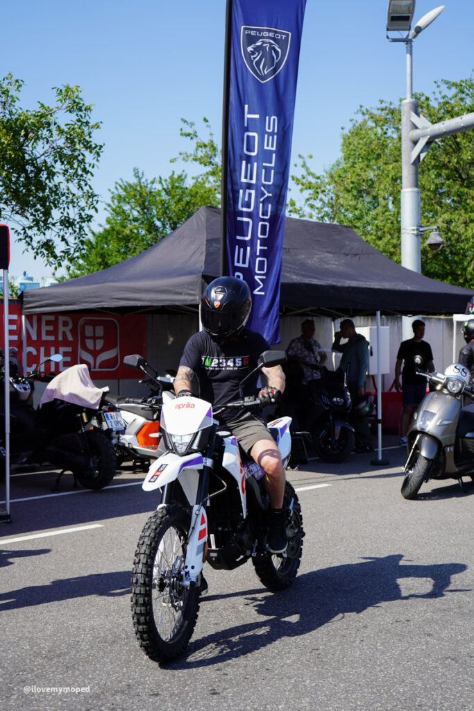 arge2rad-125ertesttag-arböfahrsicherheitszentrumwien-030 - I love my moped Motorcyclist wearing a black helmet rides a white Aprilia dirt bike at a daytime motorcycle expo, with a tall blue Peugeot banner and tents in the background.