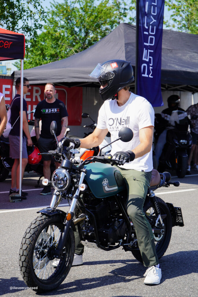 arge2rad-125ertesttag-arböfahrsicherheitszentrumwien-032 - I love my moped Motorcyclist wearing a black helmet sits on a teal retro-style bike at an outdoor event under tents.