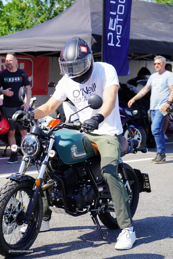 arge2rad-125ertesttag-arböfahrsicherheitszentrumwien-033 - I love my moped Man wearing a helmet sits on a teal motorcycle at an outdoor event with tents and people in the background.