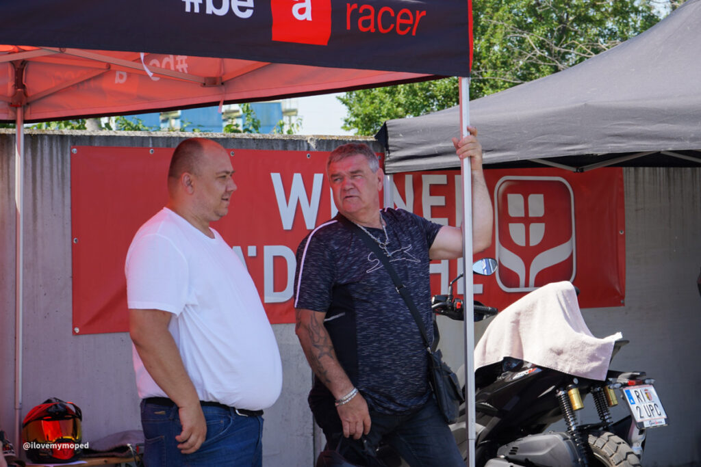 arge2rad-125ertesttag-arböfahrsicherheitszentrumwien-036 - I love my moped Two men stand under a red and black canopy, chatting beside a parked motorcycle at a street event.