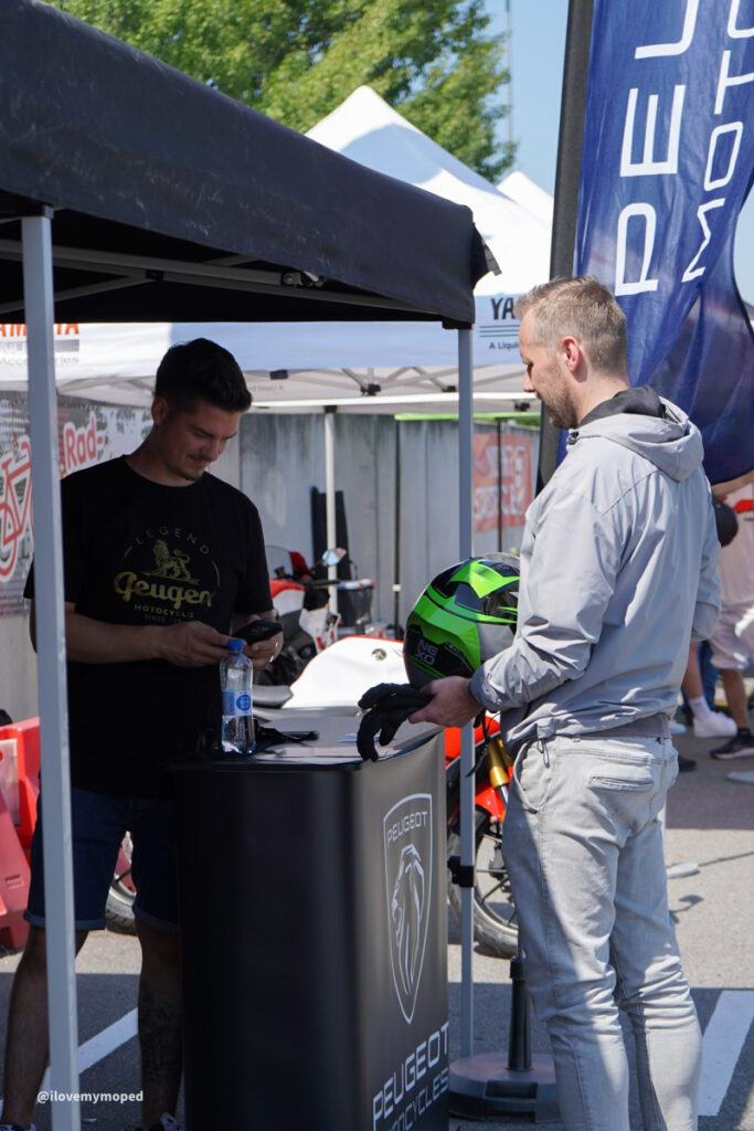 arge2rad-125ertesttag-arböfahrsicherheitszentrumwien-037 - I love my moped Man in a light gray jacket holds a bright green motorcycle helmet while talking to a booth attendant at a Peugeot motorcycles display outdoors under tents.