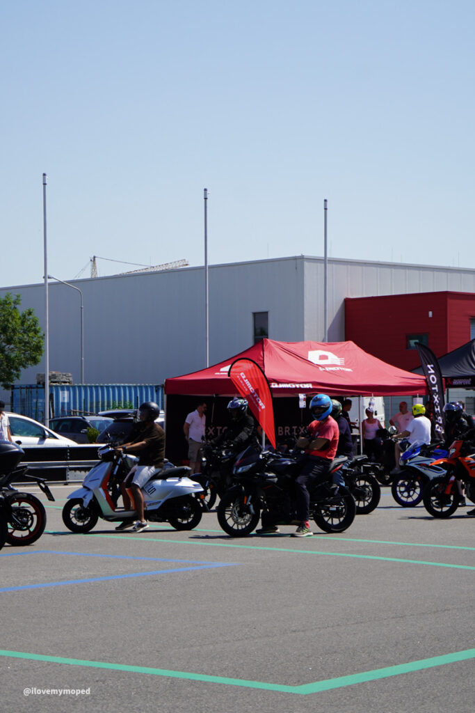 arge2rad-125ertesttag-arböfahrsicherheitszentrumwien-038 - I love my moped Motorcycle event in a parking lot with riders on scooters and sport bikes near red and black tents under a clear blue sky, vendor booths visible in the background.