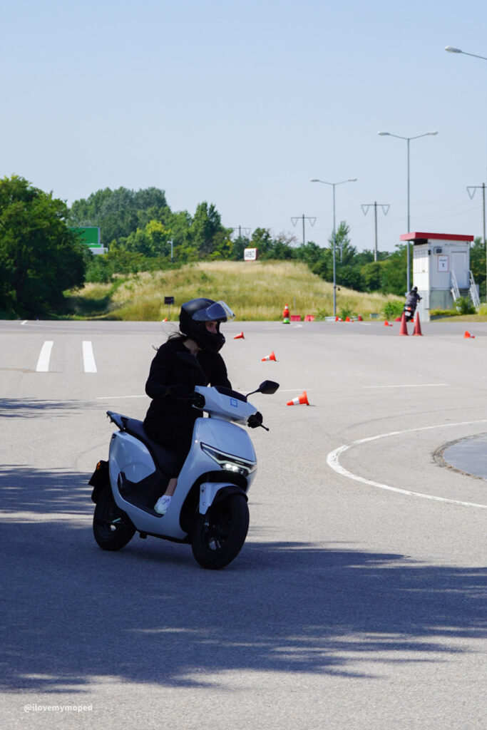 arge2rad-125ertesttag-arböfahrsicherheitszentrumwien-039 - I love my moped Person wearing a helmet rides a white scooter along a cone-marked training course on a sunny day.