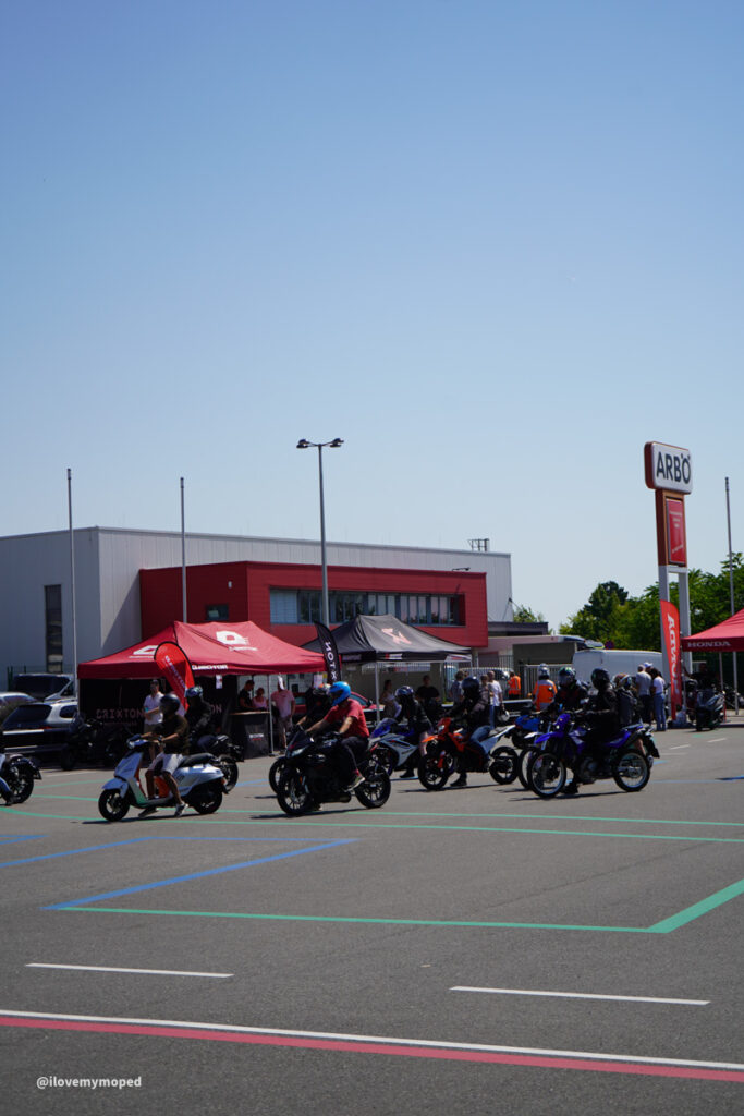 arge2rad-125ertesttag-arböfahrsicherheitszentrumwien-041 - I love my moped Motorcyclists riding in a parking lot near red tents outside a commercial building with a blue sky above.