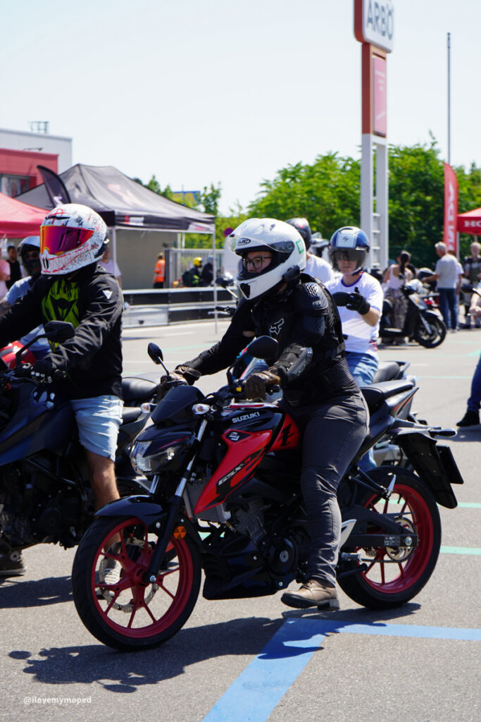 arge2rad-125ertesttag-arböfahrsicherheitszentrumwien-045 - I love my moped Motorcyclists wearing helmets prepare to ride at an outdoor event with tents in the background, Suzuki bike in front with red wheels.
