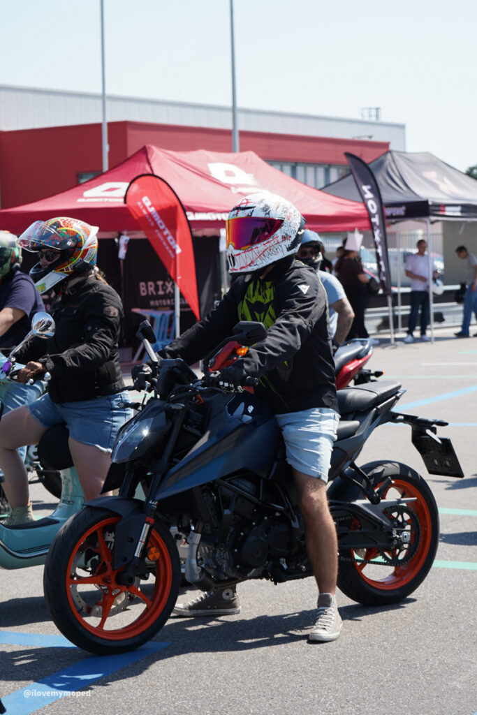 arge2rad-125ertesttag-arböfahrsicherheitszentrumwien-046 - I love my moped Motorcyclist wearing a white helmet with red visor sits on a dark blue sport bike at a bike show, red wheels visible, tents in background.