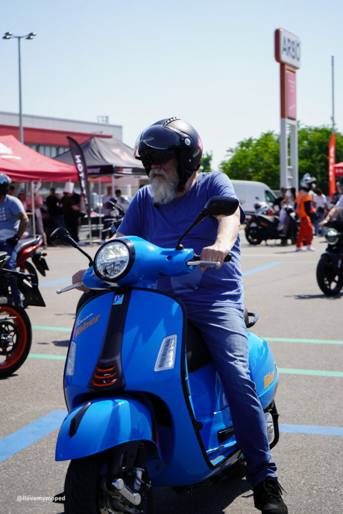 arge2rad-125ertesttag-arböfahrsicherheitszentrumwien-047 - I love my moped Older man with a gray beard wearing a black helmet rides a bright blue scooter at an outdoor motorcycle event, with tents and people in the background.