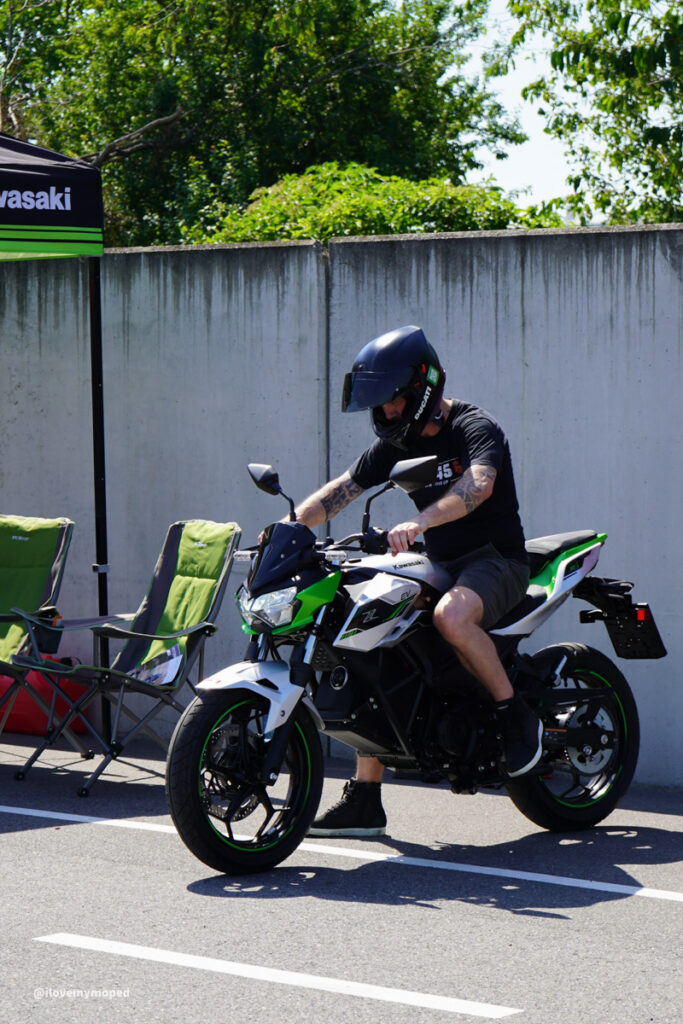 arge2rad-125ertesttag-arböfahrsicherheitszentrumwien-048 - I love my moped Man wearing a full-face helmet rides a green-and-white Kawasaki motorcycle in a parking area with green chairs nearby.