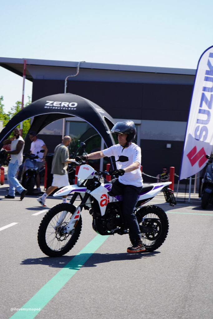 arge2rad-125ertesttag-arböfahrsicherheitszentrumwien-050 - I love my moped Rider wearing a helmet sits on a white dirt bike at an outdoor motorcycle event, with a Zero Motorcycles tent in the background.