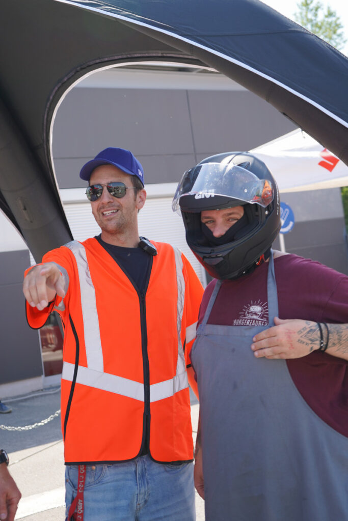 arge2rad-125ertesttag-arböfahrsicherheitszentrumwien-053 - I love my moped Man in bright orange safety vest points at something as a coworker wearing a motorcycle helmet and gray apron stands beside under a canopy at an outdoor event.