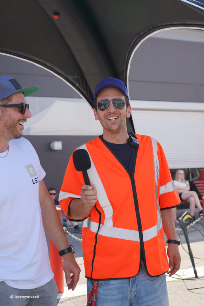 arge2rad-125ertesttag-arböfahrsicherheitszentrumwien-054 - I love my moped Man in an orange safety vest holding a microphone, being interviewed outdoors under a canopy, wearing sunglasses and a blue cap.