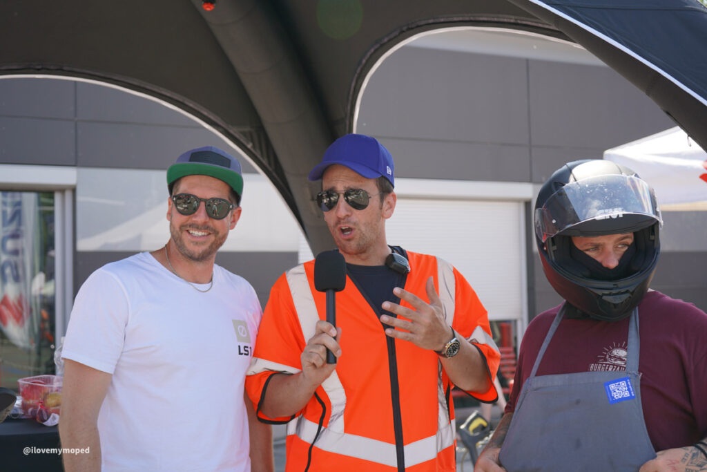 arge2rad-125ertesttag-arböfahrsicherheitszentrumwien-055 - I love my moped Three men stand under a black canopy: a smiling man in a white T-shirt and green cap wearing sunglasses, a man in an orange safety vest speaking into a microphone, and a third man in a maroon shirt with a motorcycle helmet and apron.
