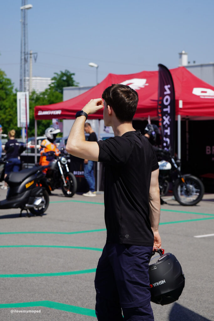 arge2rad-125ertesttag-arböfahrsicherheitszentrumwien-061 - I love my moped Man in a black t-shirt standing in a motorcycle event, holding a dark helmet at his side, with red tents and bikes in the background.