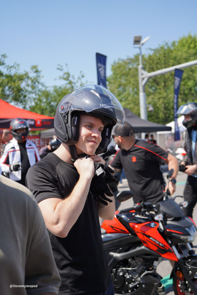 arge2rad-125ertesttag-arböfahrsicherheitszentrumwien-064 - I love my moped Person wearing a gray motorcycle helmet, adjusting the chin strap at an outdoor bike event, with a red-and-black motorcycle nearby and tents in the background.