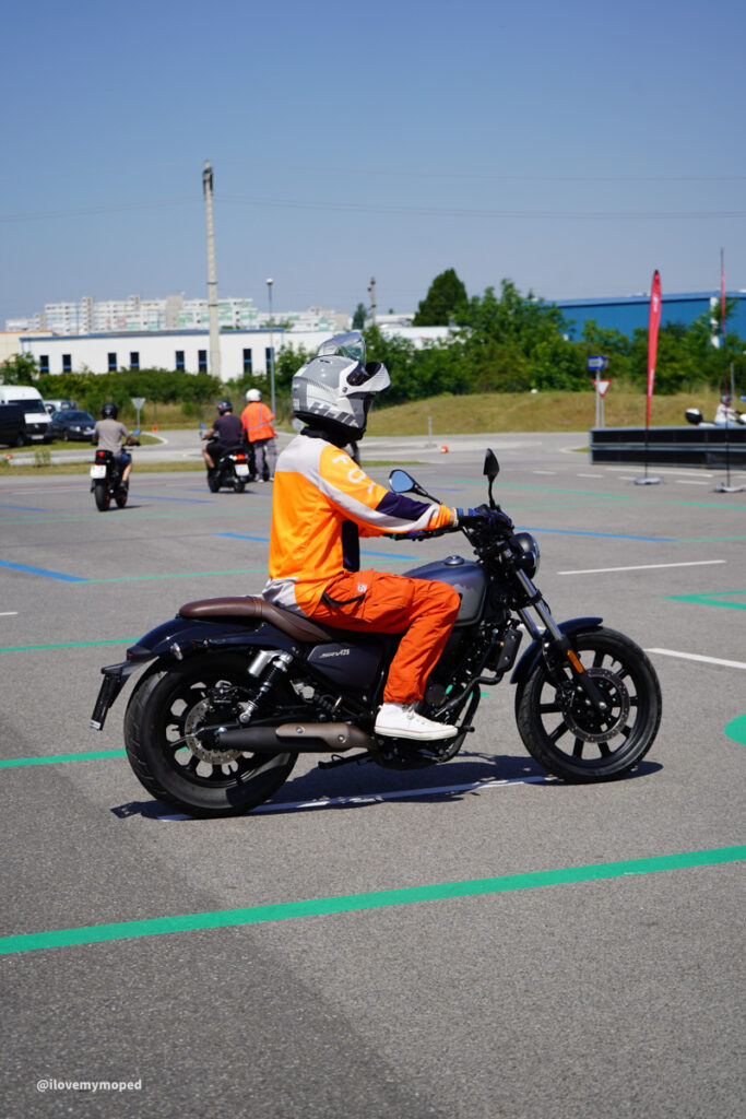 arge2rad-125ertesttag-arböfahrsicherheitszentrumwien-067 - I love my moped Motorcyclist in orange gear and white helmet riding a dark blue motorcycle in a parking lot with others in the background.