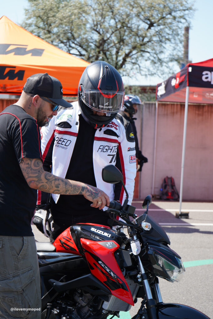 arge2rad-125ertesttag-arböfahrsicherheitszentrumwien-069 - I love my moped Rider in full-face helmet and white jacket stands beside a red Suzuki motorcycle while a man with tattoos talks to him, in a paddock area with tents in the background.