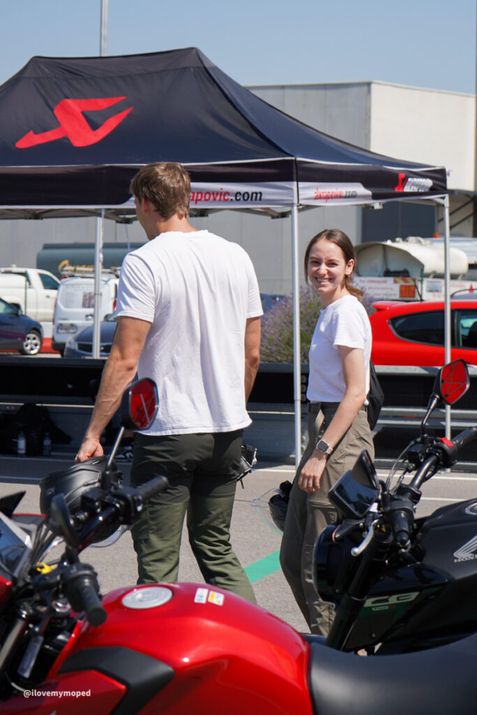 arge2rad-125ertesttag-arböfahrsicherheitszentrumwien-070 - I love my moped Man in a white t-shirt and green pants and a smiling woman in a white shirt and khaki pants stand near parked motorcycles under a black tent with a red logo.