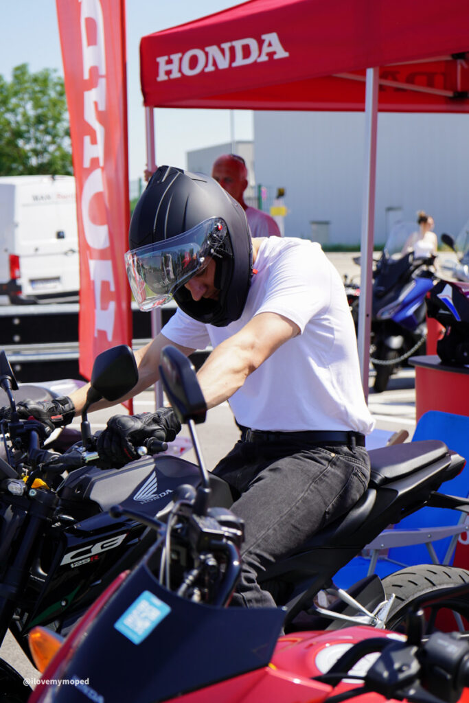 arge2rad-125ertesttag-arböfahrsicherheitszentrumwien-073 - I love my moped Person wearing a full-face helmet straddles a Honda motorcycle at an outdoor event under a red Honda tent.