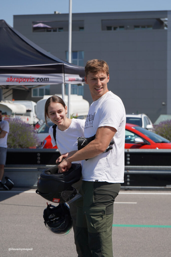 arge2rad-125ertesttag-arböfahrsicherheitszentrumwien-074 - I love my moped Two people at an outdoor motorsport event; a man holds a motorcycle helmet while a smiling woman stands beside him under an akrapovic tent.
