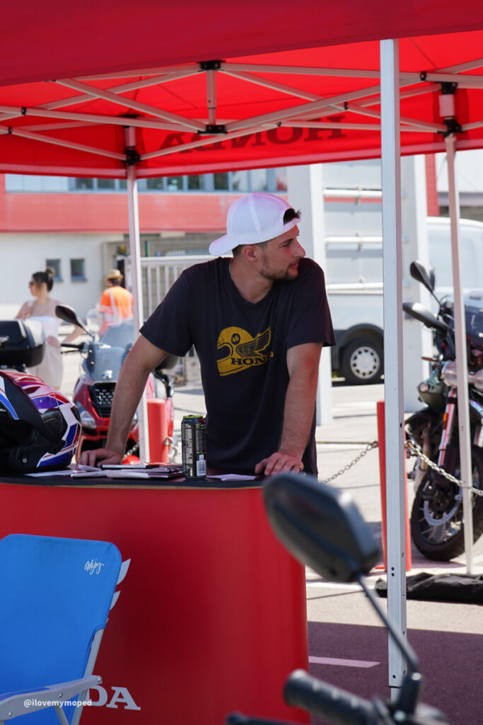 arge2rad-125ertesttag-arböfahrsicherheitszentrumwien-075 - I love my moped Man in a white backward cap and black Honda t-shirt leans on a red booth under a red canopy at a motorcycle event, with bikes and a helmet nearby.