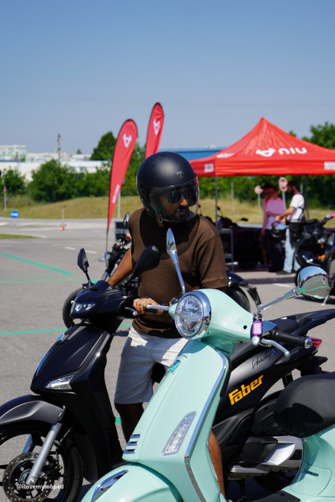 arge2rad-125ertesttag-arböfahrsicherheitszentrumwien-076 - I love my moped Man wearing a helmet and sunglasses sits on a mint-green Vespa scooter at an outdoor event with red tents in the background.