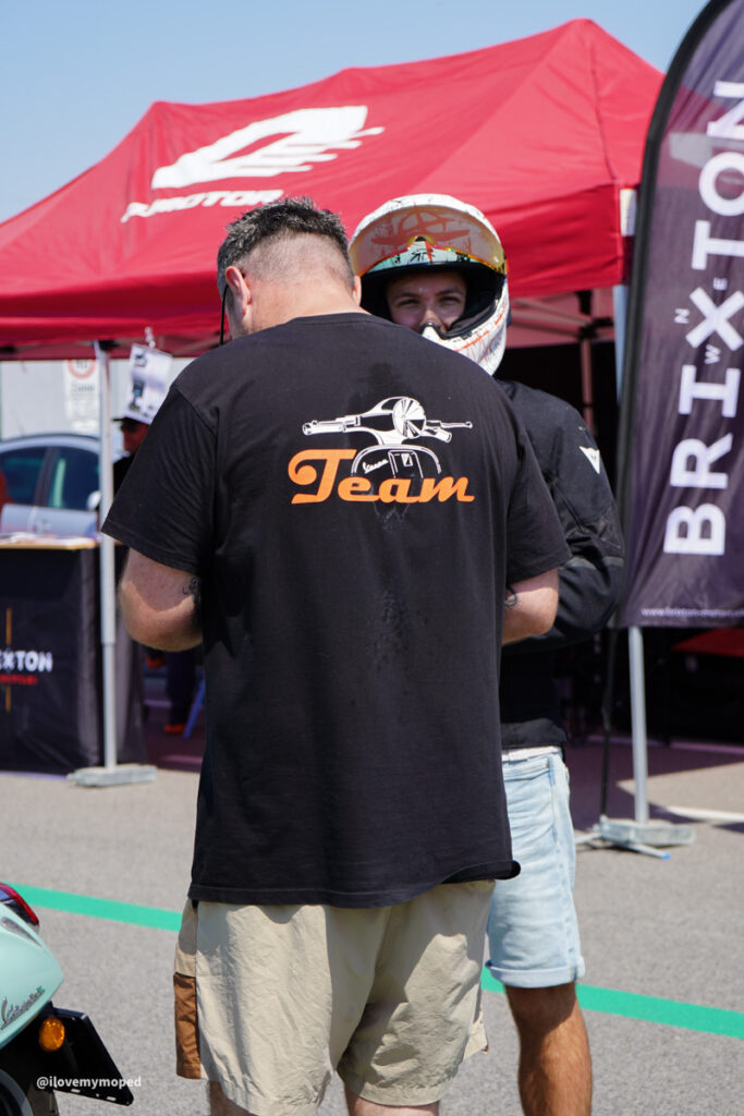 arge2rad-125ertesttag-arböfahrsicherheitszentrumwien-077 - I love my moped Two people at an outdoor event near a red tent; one man wears a black 'Team' T‑shirt with an aircraft graphic, facing away from the camera, the other in a helmet.