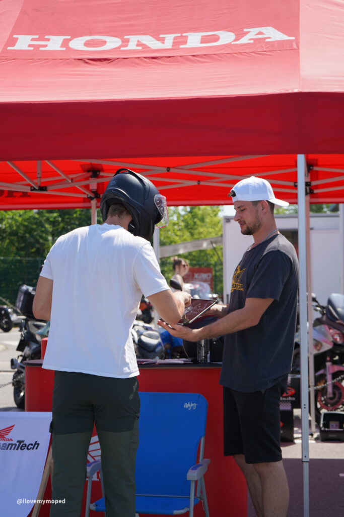 arge2rad-125ertesttag-arböfahrsicherheitszentrumwien-079 - I love my moped Two men under a red Honda tent at a motorcycle event; one in a white shirt and helmet hands something to the other man in a dark shirt and white cap, with motorcycles in the background.