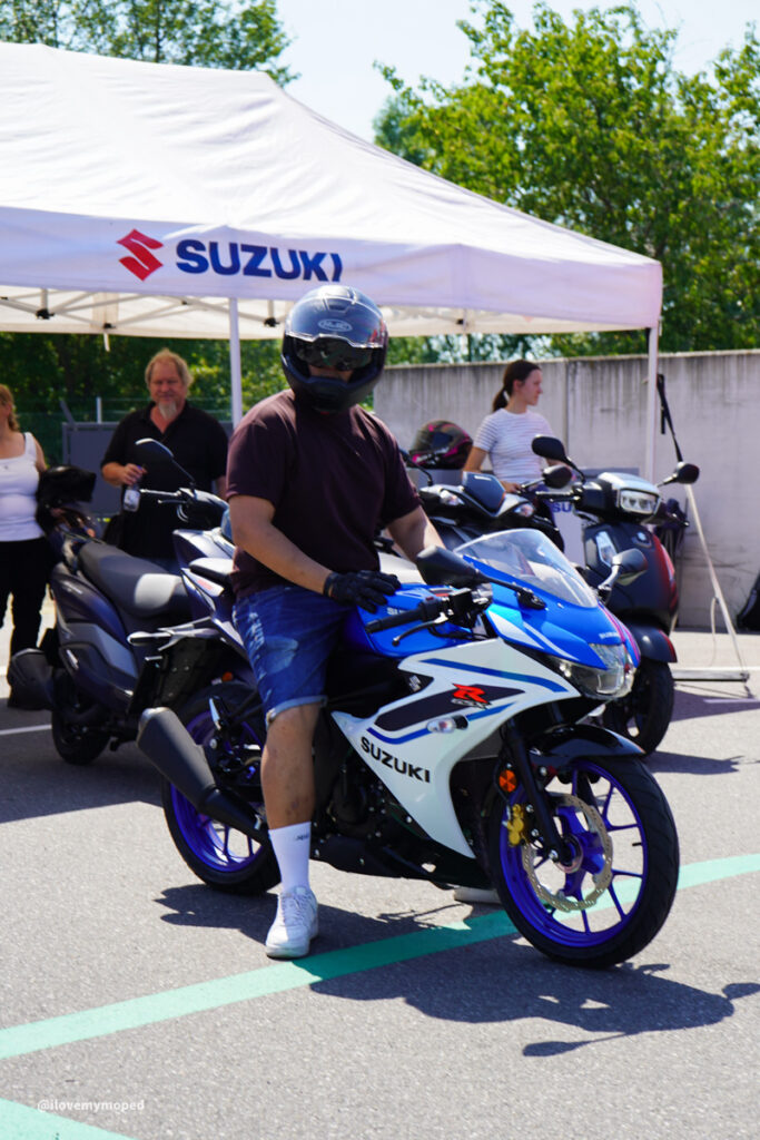 arge2rad-125ertesttag-arböfahrsicherheitszentrumwien-081 - I love my moped Rider wearing helmet and maroon shirt sits on a blue-and-white Suzuki GSX-R sport bike at an outdoor Suzuki event under a white tent with onlookers in the background.