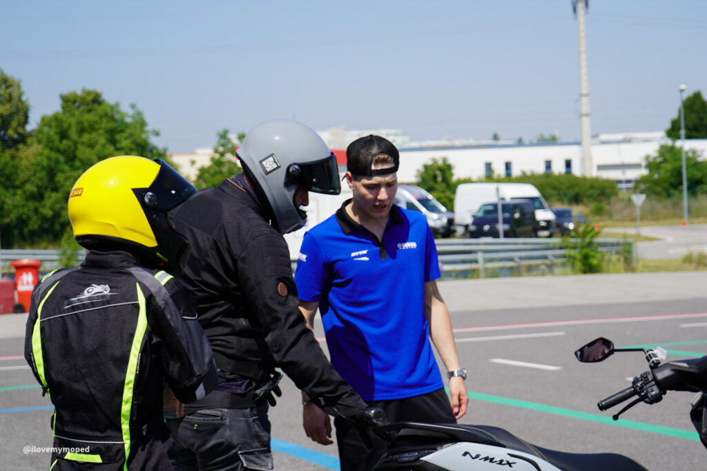 arge2rad-125ertesttag-arböfahrsicherheitszentrumwien-082 - I love my moped Two motorcyclists in gear and a man in a blue polo discuss a white motorcycle in a parking lot.