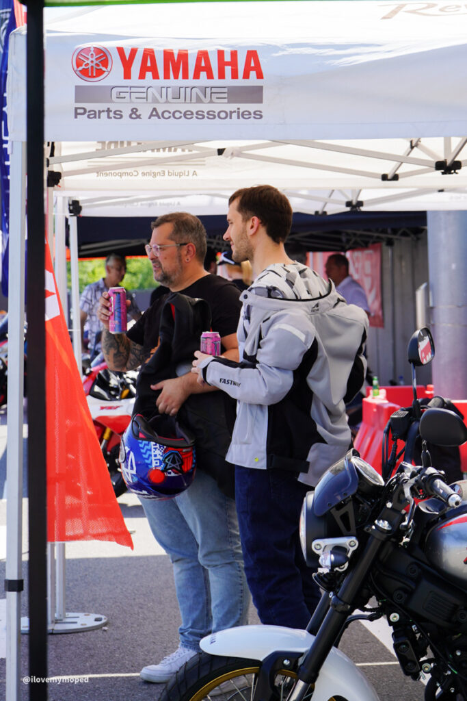 arge2rad-125ertesttag-arböfahrsicherheitszentrumwien-086 - I love my moped Two men stand under a Yamaha Genuine Parts & Accessories tent, holding pink cans, with a motorcycle in the foreground.
