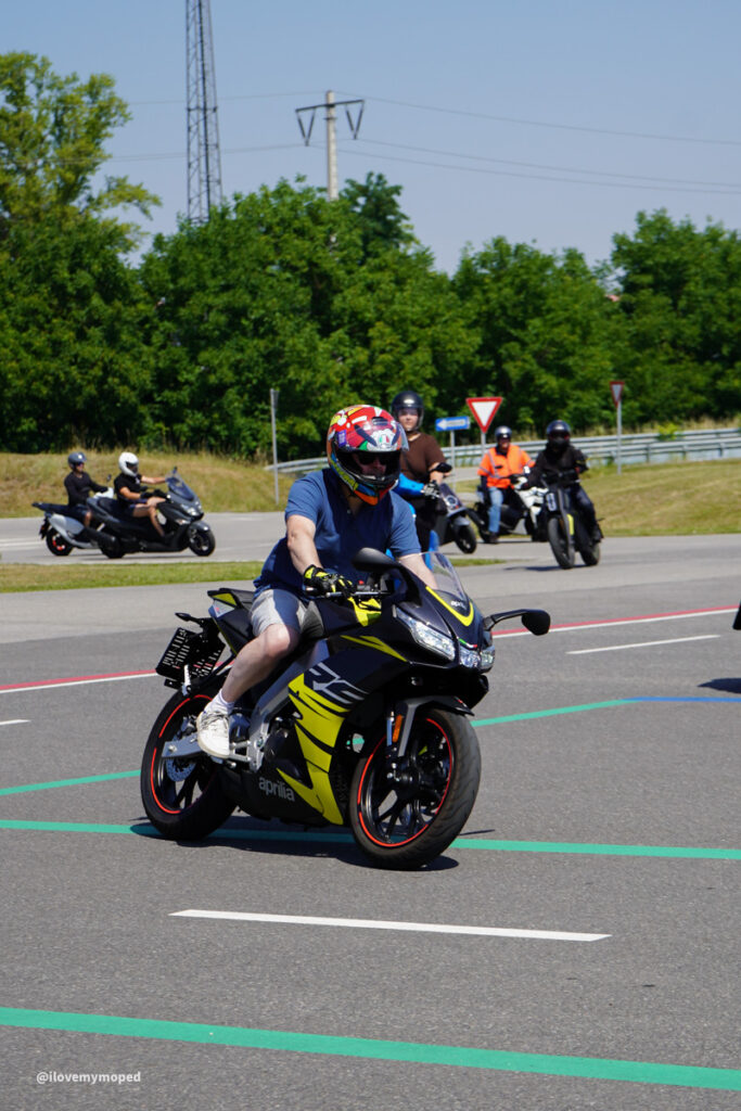 arge2rad-125ertesttag-arböfahrsicherheitszentrumwien-087 - I love my moped Rider in a blue shirt wearing a colorful full-face helmet on a black and yellow Aprilia sportbike turning on a paved course, with other riders and spectators in the background.