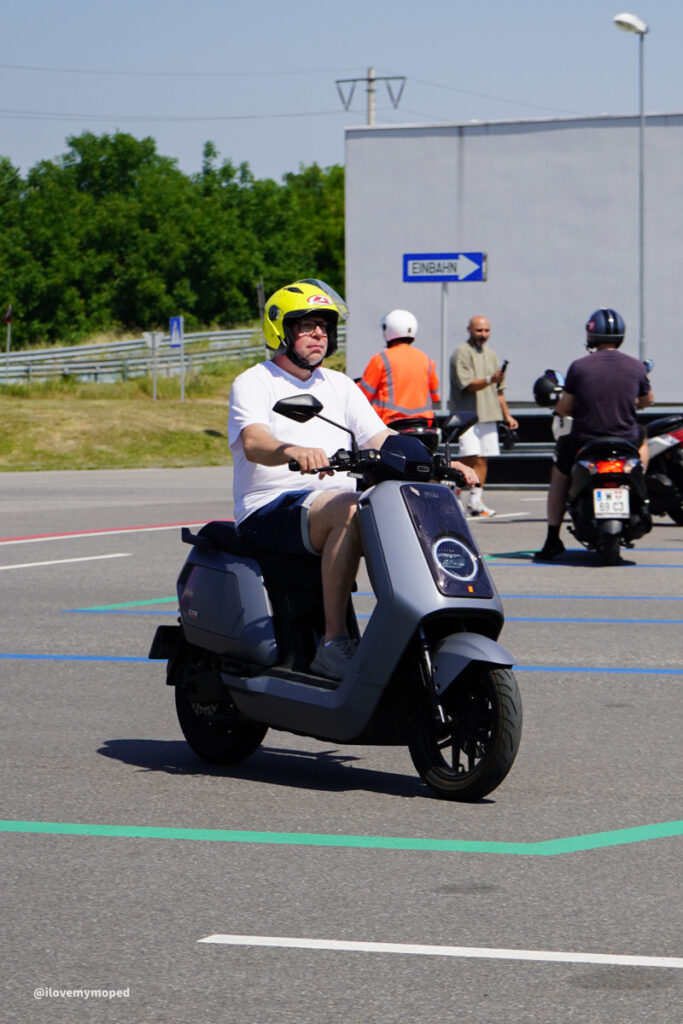 arge2rad-125ertesttag-arböfahrsicherheitszentrumwien-088 - I love my moped Man wearing a yellow helmet rides a gray scooter in a parking lot or training area with other riders in the background.