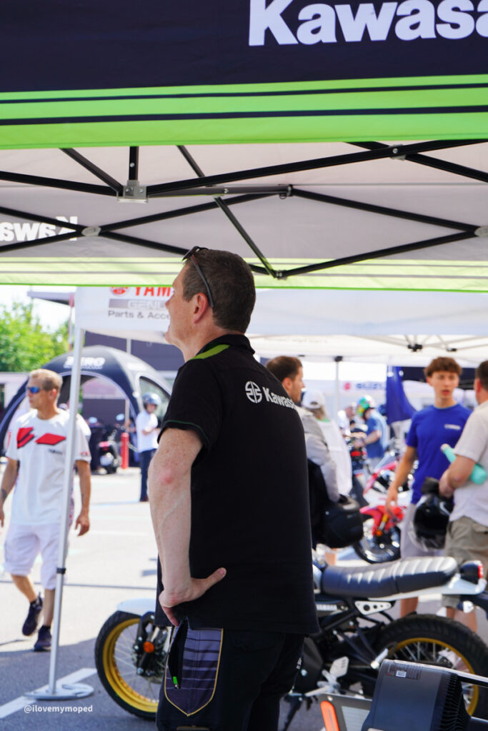 arge2rad-125ertesttag-arböfahrsicherheitszentrumwien-089 - I love my moped Man in a black Kawasaki polo stands under a green-and-black tent at a motorcycle event, with a custom bike nearby.