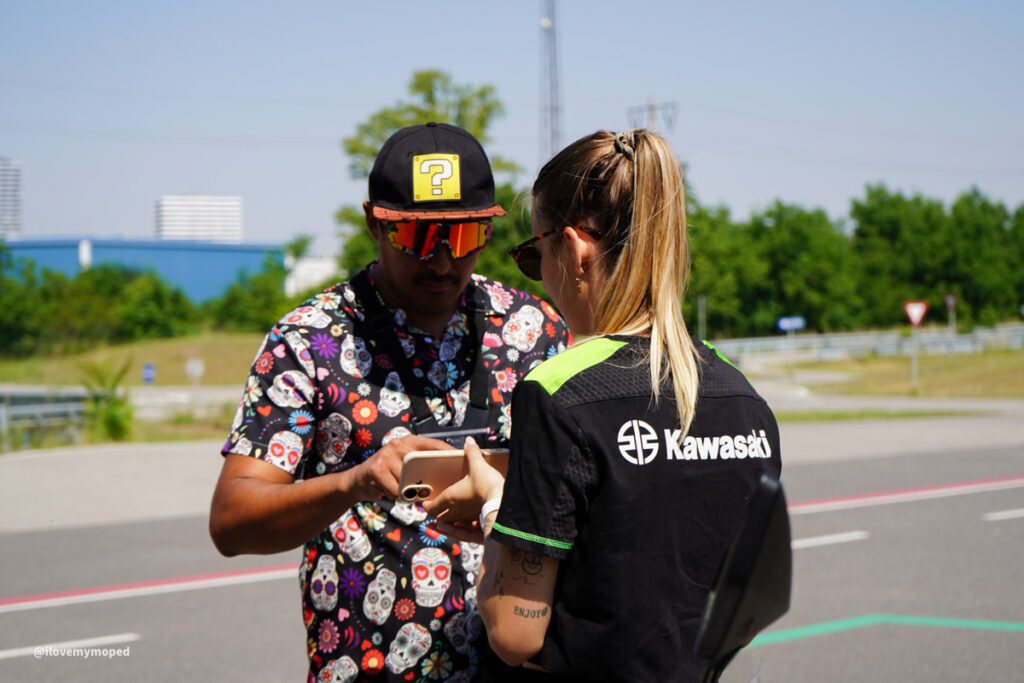 arge2rad-125ertesttag-arböfahrsicherheitszentrumwien-092 - I love my moped Man wearing a skull-pattern shirt and a cap with a question mark shows a smartphone to a woman in a Kawasaki shirt outdoors on a sunny day.