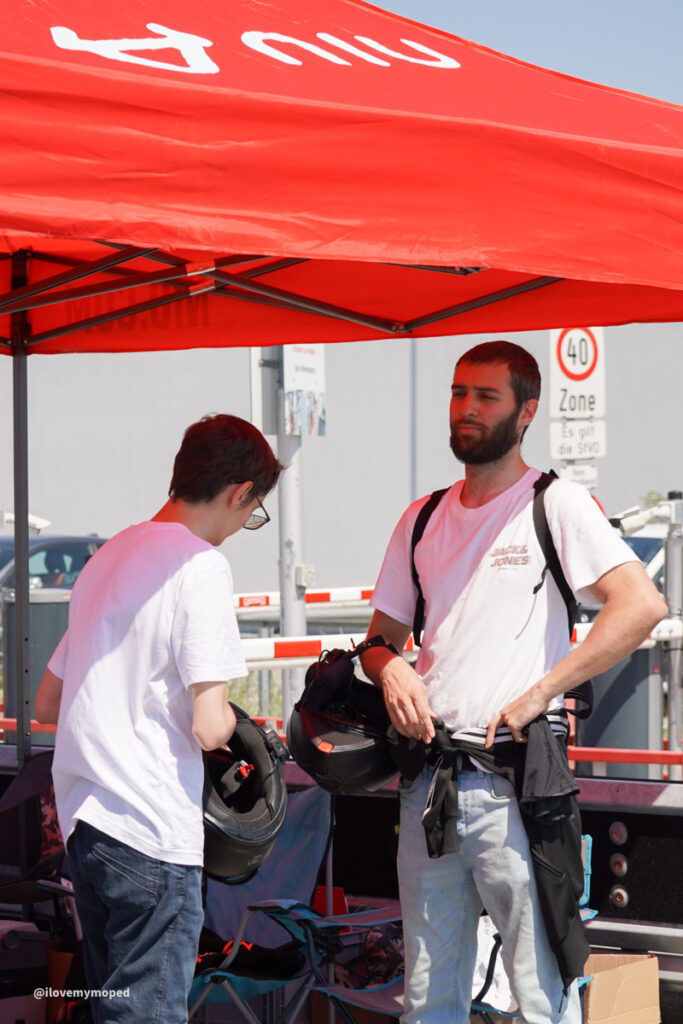 arge2rad-125ertesttag-arböfahrsicherheitszentrumwien-093 - I love my moped Two men in white T-shirts under a red canopy, one handing a black motorcycle helmet to the other in a parking lot setting with gear nearby: