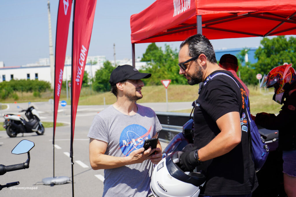 arge2rad-125ertesttag-arböfahrsicherheitszentrumwien-095 - I love my moped Two men chat under a red tent at a motorcycle event; one wears a NASA shirt and holds a phone.