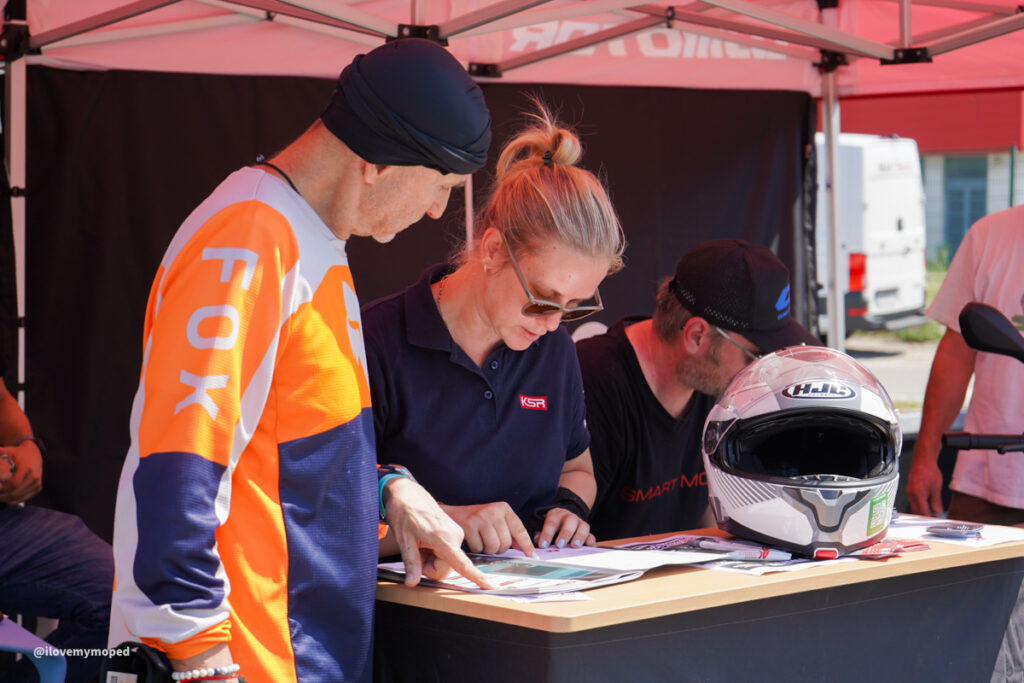 arge2rad-125ertesttag-arböfahrsicherheitszentrumwien-096 - I love my moped Two people sign documents at a table under a red canopy, with a full-face motorcycle helmet nearby on the desk.