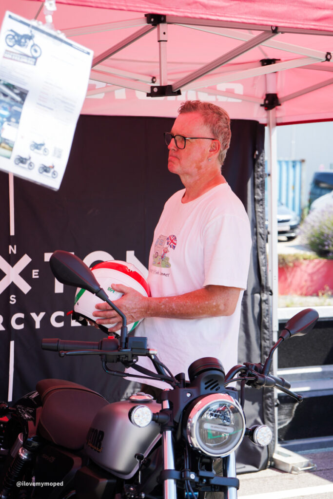 arge2rad-125ertesttag-arböfahrsicherheitszentrumwien-097 - I love my moped Man with short gray hair and glasses stands beside a silver motorcycle under a pink canopy, holding a white helmet with red accents.
