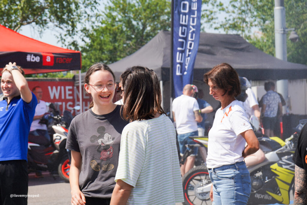 arge2rad-125ertesttag-arböfahrsicherheitszentrumwien-100 - I love my moped Group of people chatting and smiling at a daytime outdoor motorcycling event with tents and bikes in the background.