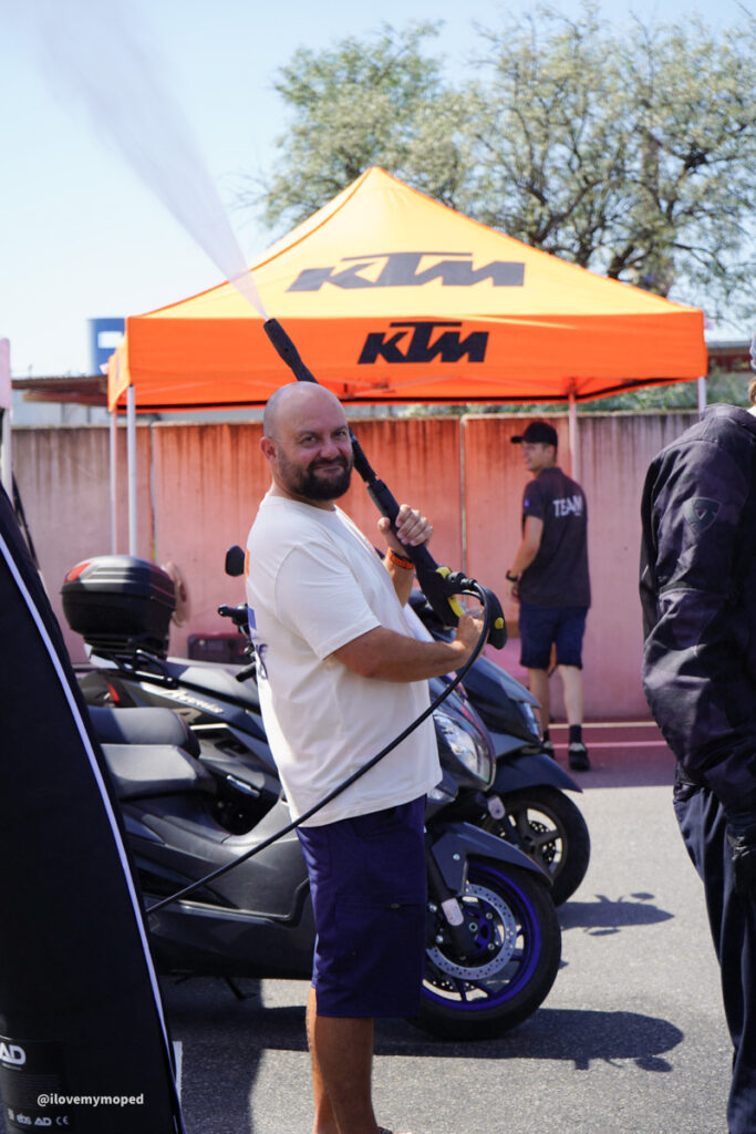 arge2rad-125ertesttag-arböfahrsicherheitszentrumwien-101 - I love my moped Man in a white t-shirt and blue shorts holding a large spray nozzle at a KTM booth with orange canopy, standing beside a blue motorcycle.