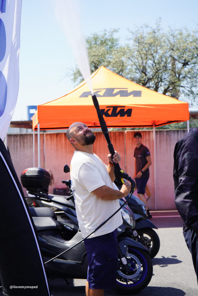 arge2rad-125ertesttag-arböfahrsicherheitszentrumwien-102 - I love my moped Man in a white t-shirt and blue shorts holds a long black hose with a nozzle at a motorcycle event under an orange KTM tent, with bikes in the background.