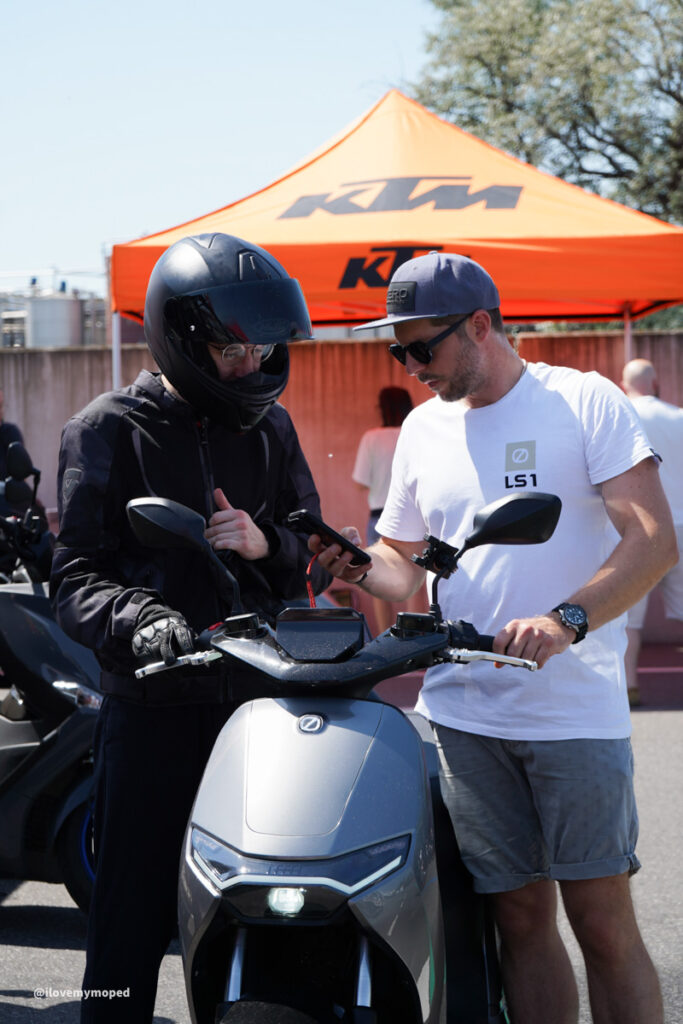arge2rad-125ertesttag-arböfahrsicherheitszentrumwien-103 - I love my moped Man in a full-face helmet and black riding gear shows a phone to another man in a white T-shirt and cap beside a gray scooter under an orange KTM tent.